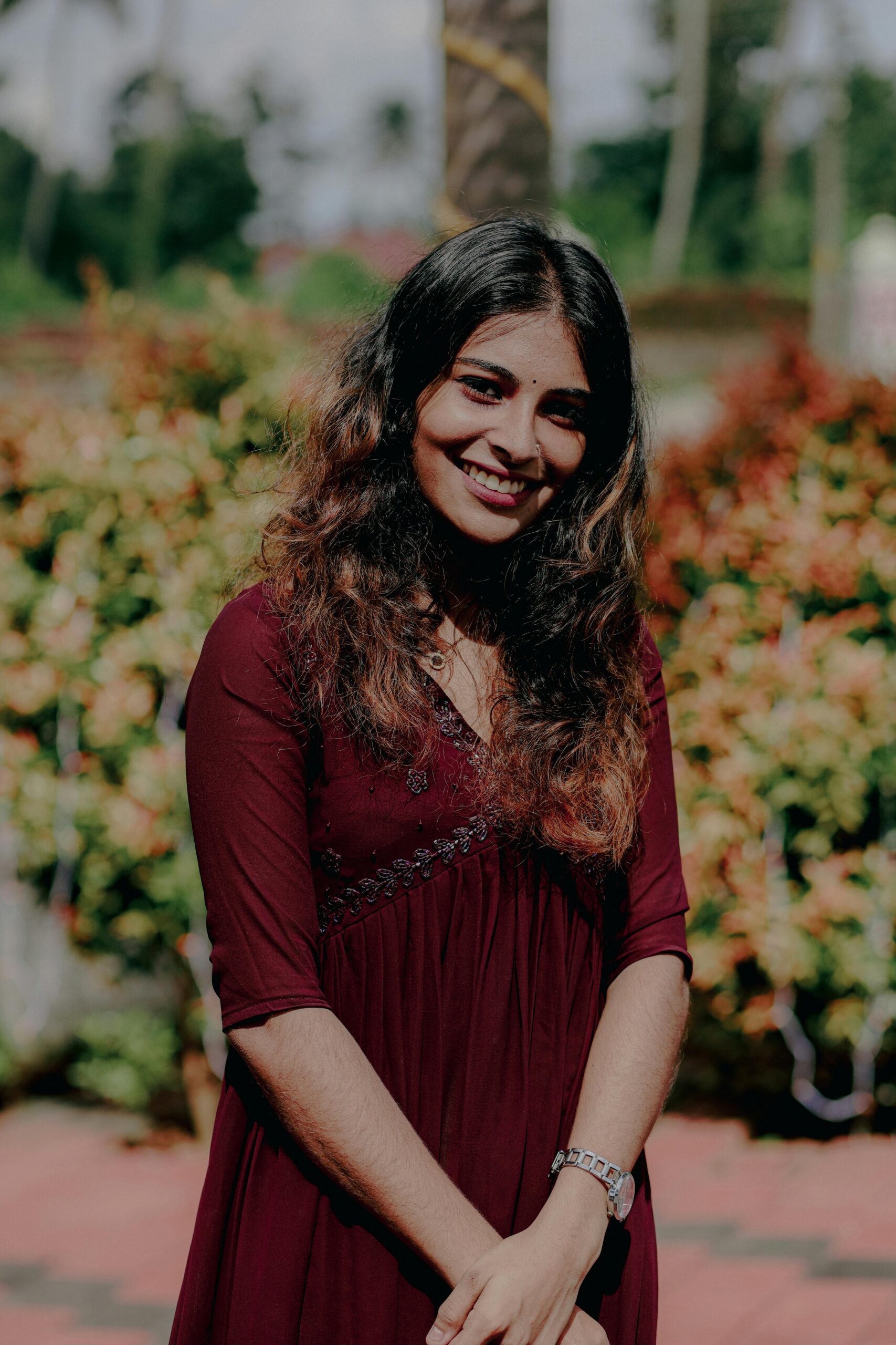 Portrait of a smiling woman in a maroon dress standing outdoors with lush greenery in Kochi, India.