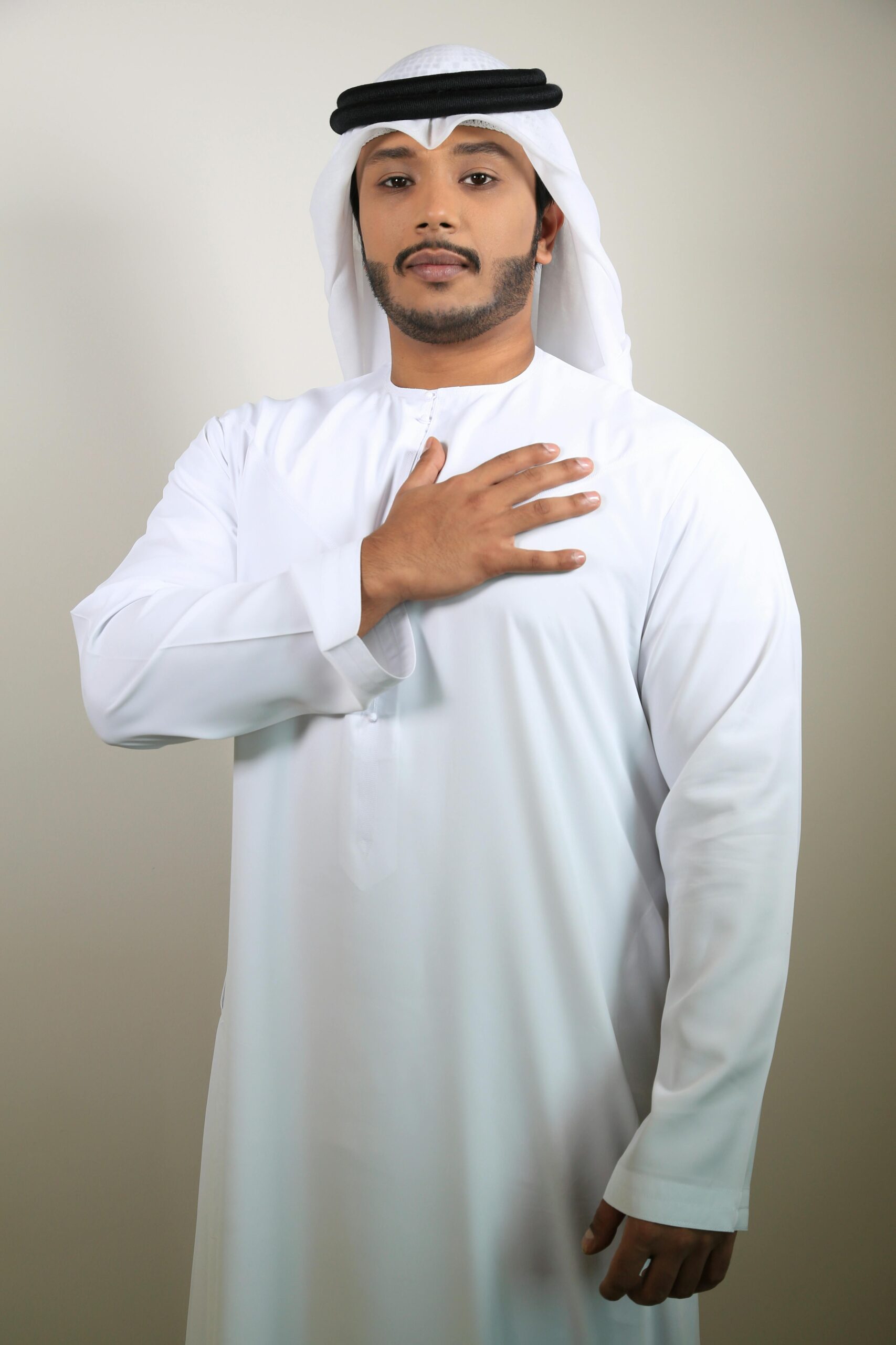 Portrait of an adult man in traditional Emirati clothing with hand on chest against plain background.