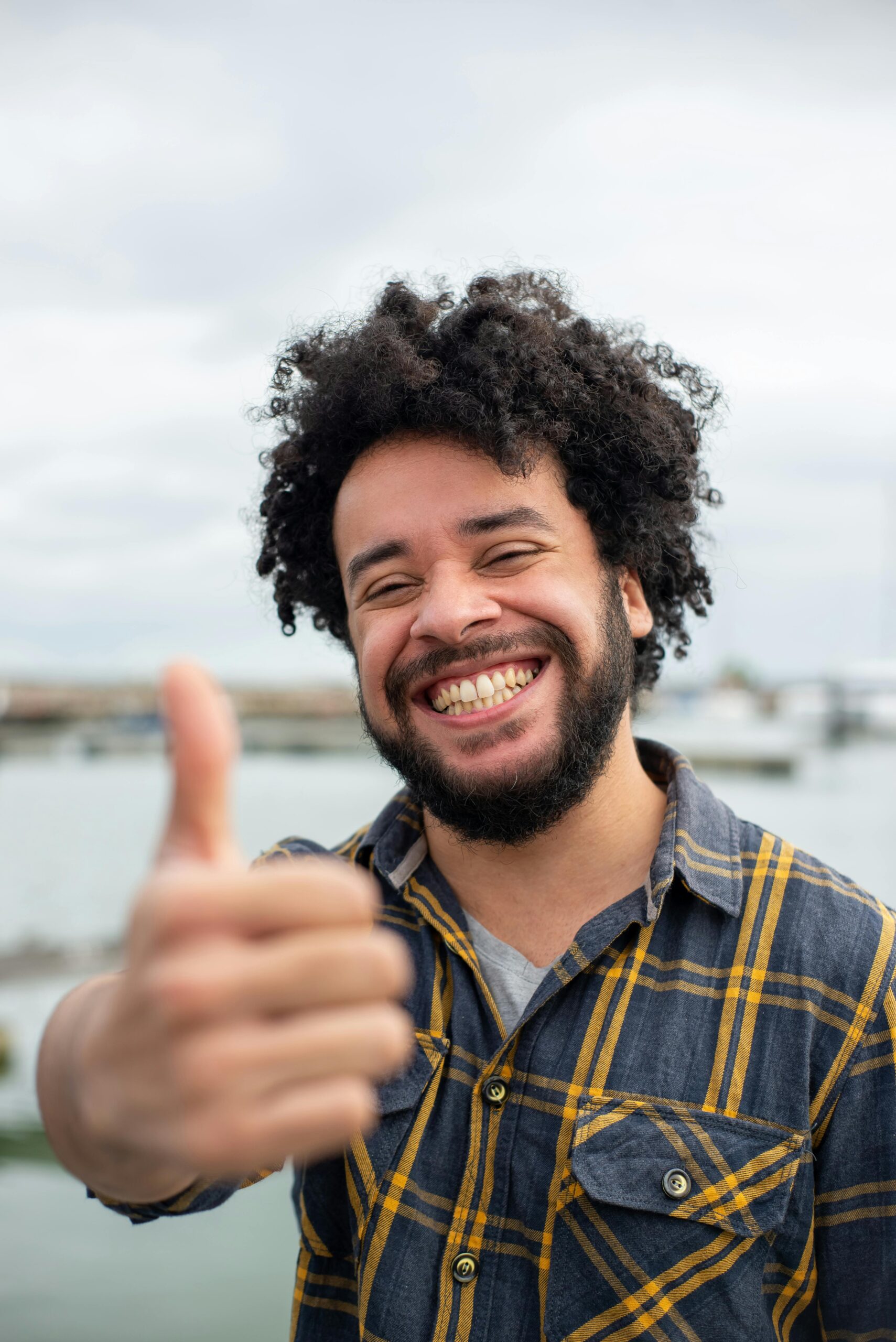 Cheerful man with afro hair and beard smiling, giving thumbs up in scenic outdoor setting.